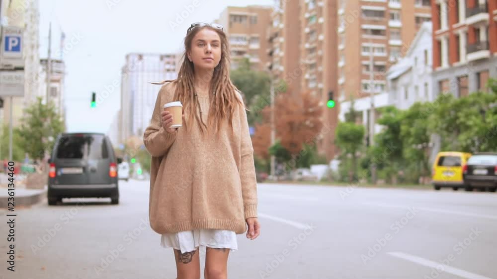 young caucasian woman walking along the road in urban city slow motion. caucasian model with dreadlocks wearing oversized sweater holding cup of coffee drinking on the run outdoors young caucasian woman walking along the road in urban city slow motion. caucasian model with dreadlocks wearing oversized sweater holding cup of coffee drinking on the run outdoors