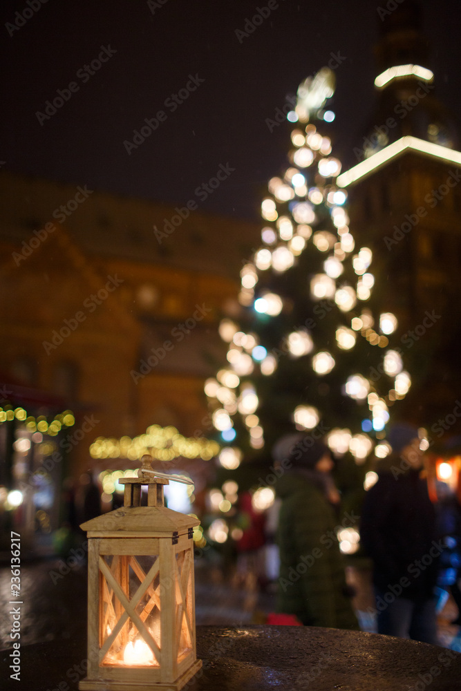 a lantern with a candle on the background of a city fair and a Christmas tree with lights