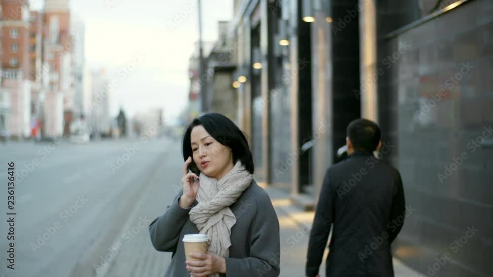 Tilt down shot of middle-aged Asian woman holding takeaway coffee cup and talking on the phone while standing on pavement Tilt down shot of middle-aged Asian woman holding takeaway coffee cup and talking on the phone while standing on pavement