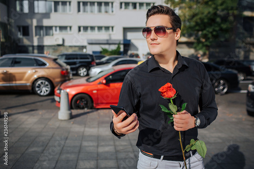 Happy young man stand and smile. He hold phone and red rose in hands. There are road with cars behind. It is sunny outside.