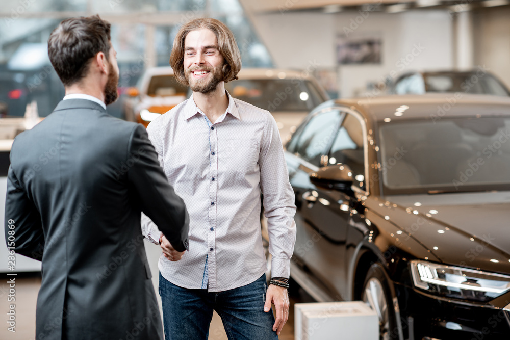 Young man shaking hand with professional salesperson having a deal in the showroom with luxury cars on the background