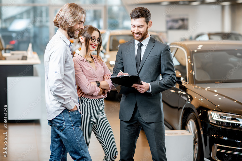 Obraz premium Handsome male dealer selling to a young couple clients a new car, standing together with some documents in the showroom