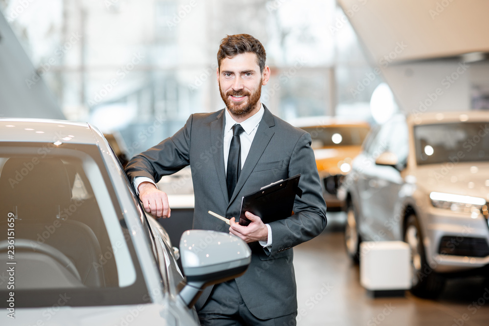 Portrait of a handsome salesman in the suit standing near the car in the showroom Portrait of a handsome salesman in the suit standing near the car in the showroom