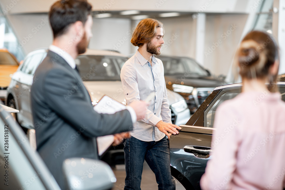 Obraz premium Young couple with salesperson choosing a new car looking on the back seats in the showroom