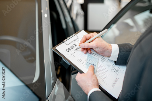 Manager filling car rental documents holding folder with driver license in the showroom with car on the background