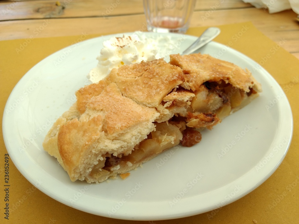 A strudel cake slice with some whipped cream, apples and raisin on a white dish with a spoon