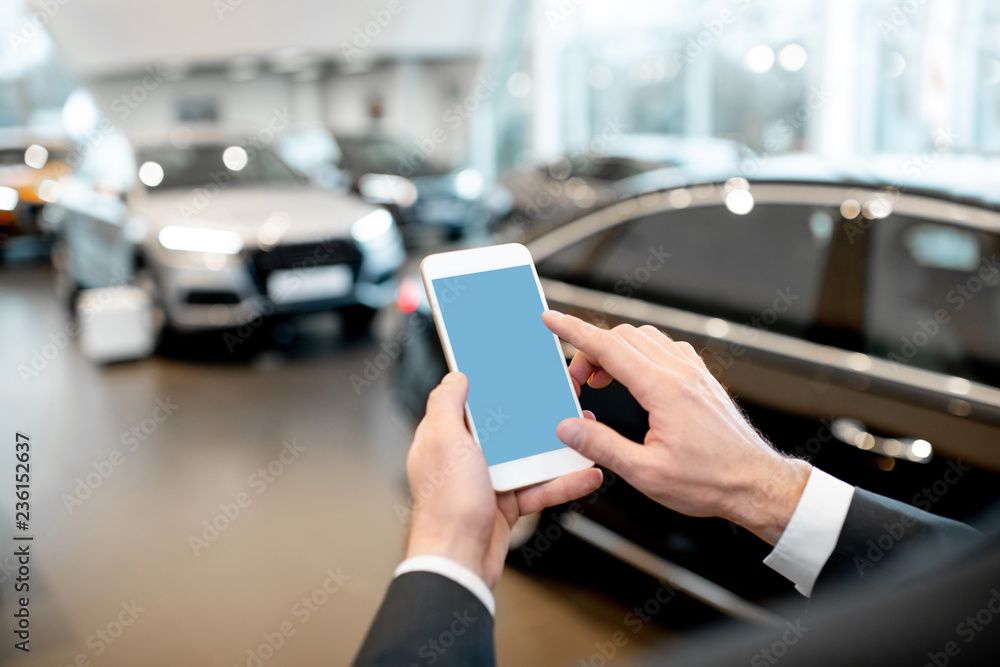 Businessman holding phone with empty screen to copy space in the showroom with cars on the background