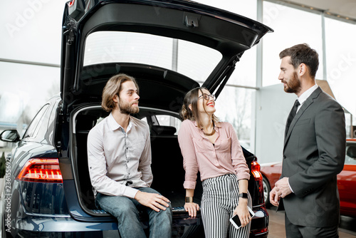 Young couple sitting in the trunk of a new car listening to the salesman at the showroom