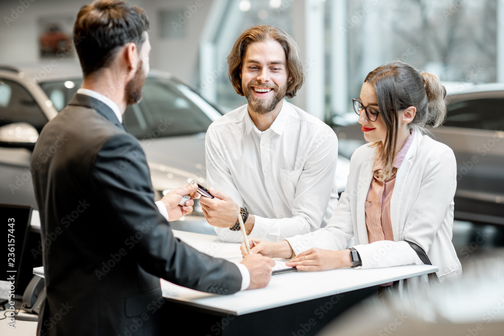 Young business couple signing some documents at the table with salesperson or manager buying or renting car in the showroom Young business couple signing some documents at the table with salesperson or manager buying or renting car in the showroom