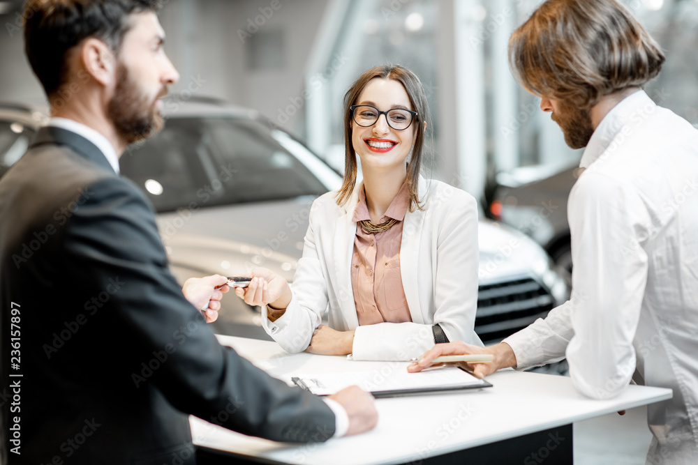 Young business couple signing some documents at the table with salesperson or manager buying or renting car in the showroom Young business couple signing some documents at the table with salesperson or manager buying or renting car in the showroom