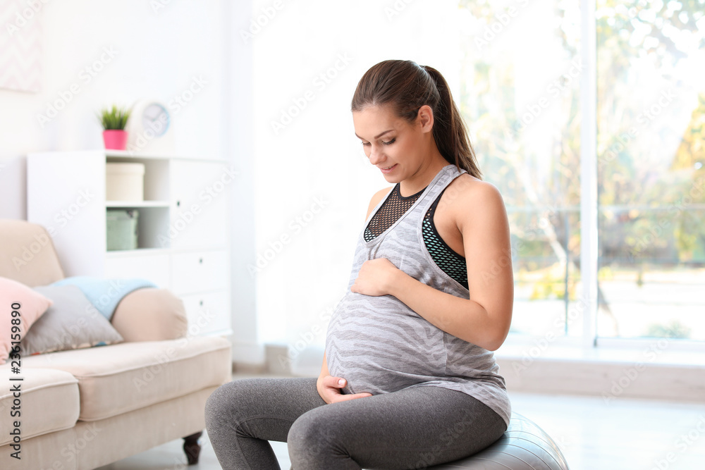 Young pregnant woman in fitness clothes sitting on exercise ball at home