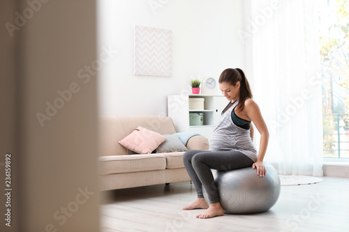 Young pregnant woman in fitness clothes sitting on exercise ball at home. Space for text