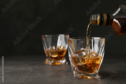 Fototapeta Naklejka Na Ścianę i Meble -  Pouring whiskey from bottle into glass with ice cubes on table. Space for text