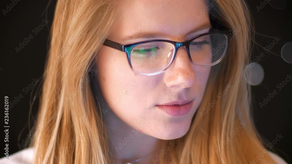 Concentrated face close-up, blonde in spectacles is looking at phone screen while standing , green screen reflected in glasses, dark background, smart person Concentrated face close-up, blonde in spectacles is looking at phone screen while standing , green screen reflected in glasses, dark background, smart person