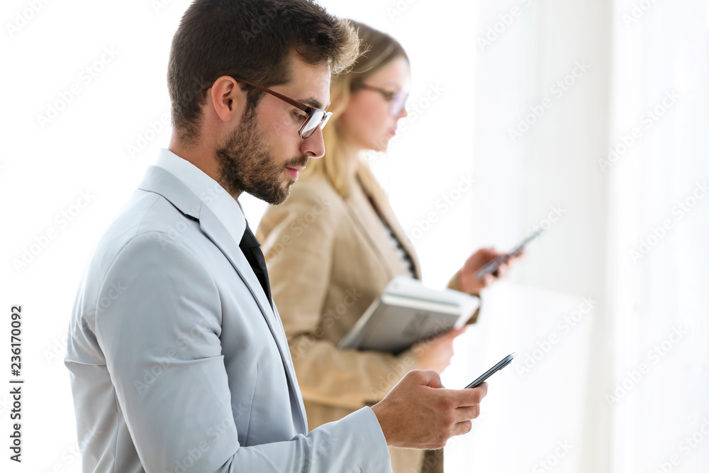 Two attractive young business partners texting with their smartphones in a hallway of they company.