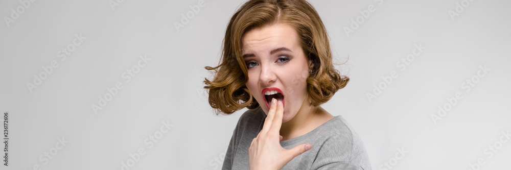 Charming young girl in a gray T-shirt on a gray background. The girl put two fingers in her mouth Charming young girl in a gray T-shirt on a gray background. The girl put two fingers in her mouth
