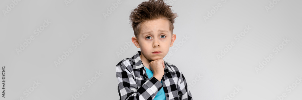 A handsome boy in a plaid shirt, blue shirt and jeans stands on a gray background. The boy supports his chin with his hand A handsome boy in a plaid shirt, blue shirt and jeans stands on a gray background. The boy supports his chin with his hand