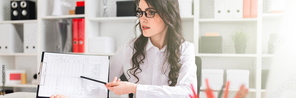 A beautiful young girl is sitting at a table in the office and pointing a pencil at the information in the document. A beautiful young girl is sitting at a table in the office and pointing a pencil at the information in the document.