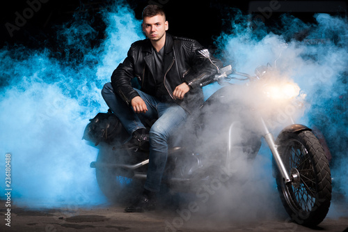Cute biker in leather jacket sits on a motorcycle in blue smoke