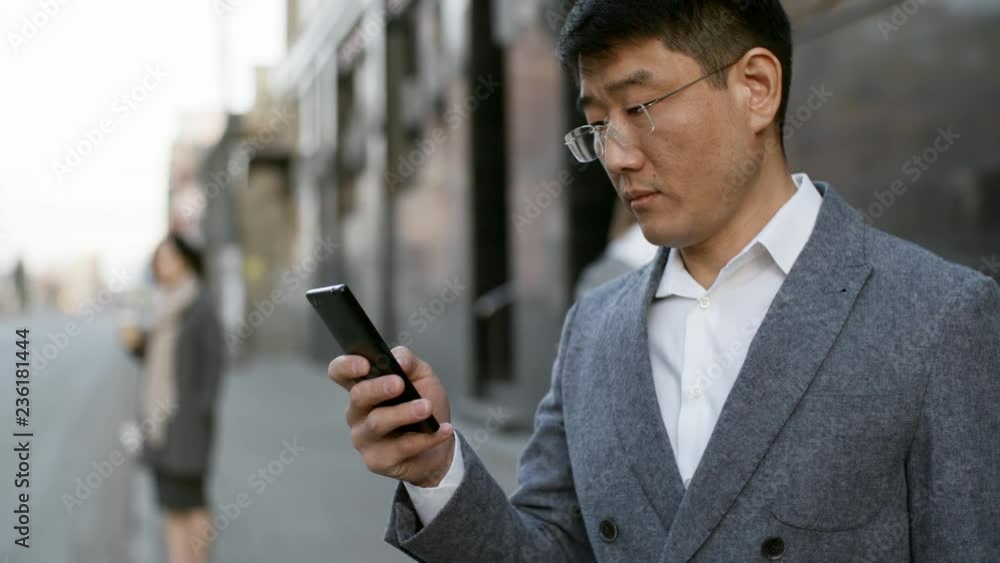 Tracking medium shot of middle-aged Asian man in glasses standing on pavement and using smartphone while waiting for transport at bus stop Tracking medium shot of middle-aged Asian man in glasses standing on pavement and using smartphone while waiting for transport at bus stop