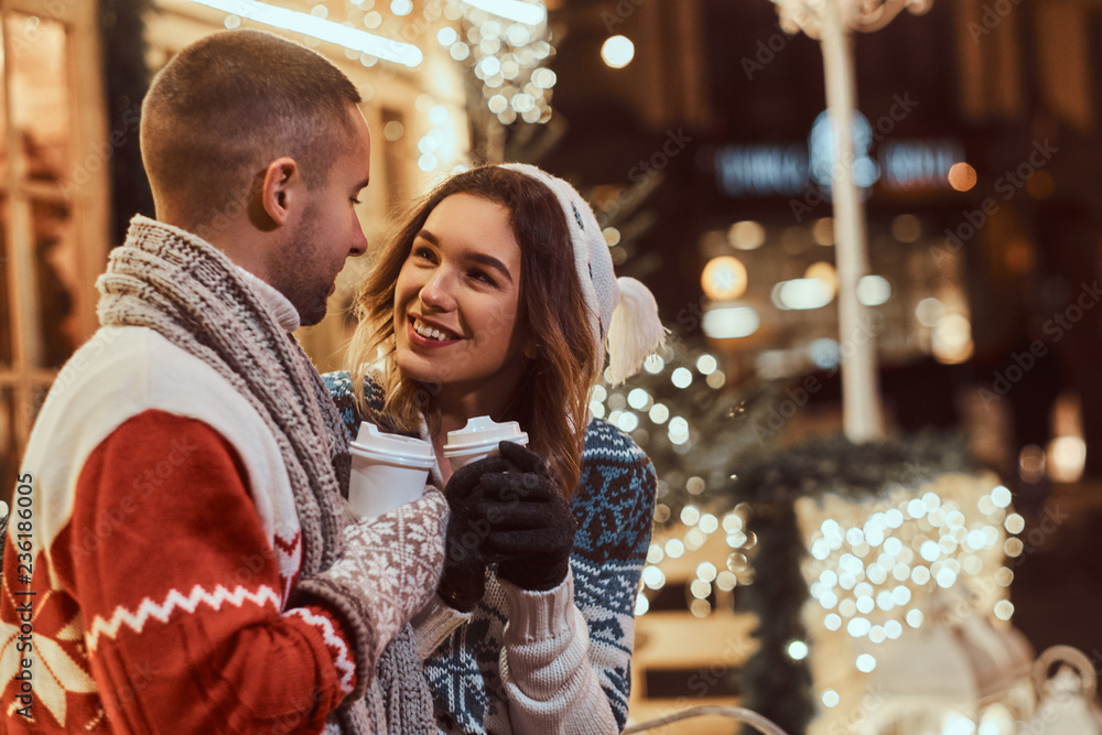 A young romantic couple wearing warm clothes outdoor at Christmas time, sitting on a bench and warming with hot coffee in evening street decorated with beautiful lights. A young romantic couple wearing warm clothes outdoor at Christmas time, sitting on a bench and warming with hot coffee in evening street decorated with beautiful lights.