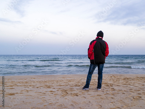 A lone man is standing on an empty sea beach and looking into the distance on a cold overcast autumn day, view from the back. Loneliness concept