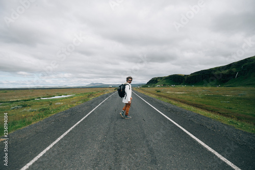 Trendy hipster millennial young man with funny curly hair walks in middle of empty road or highway, looks back at camera, urban nomad travels path less explored durign roadtrip in iceland