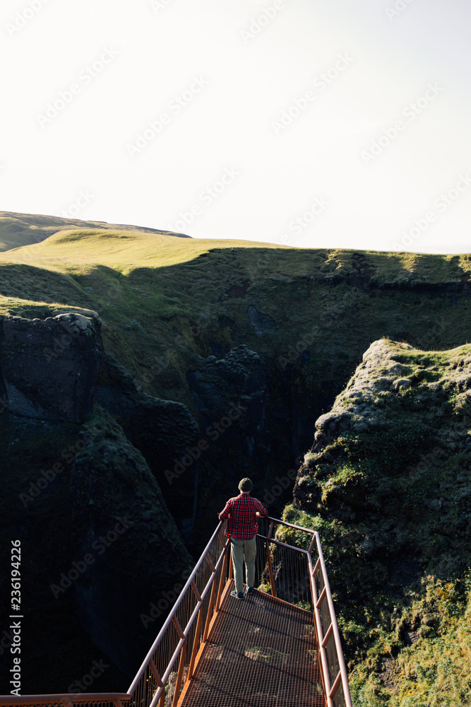Tourist or traveler man in red checkered shirt and beanie hat stands on viewing point overlooking inspiring epic mountain valley at sunset, new experiences and adrenaline emotions for adventures Tourist or traveler man in red checkered shirt and beanie hat stands on viewing point overlooking inspiring epic mountain valley at sunset, new experiences and adrenaline emotions for adventures