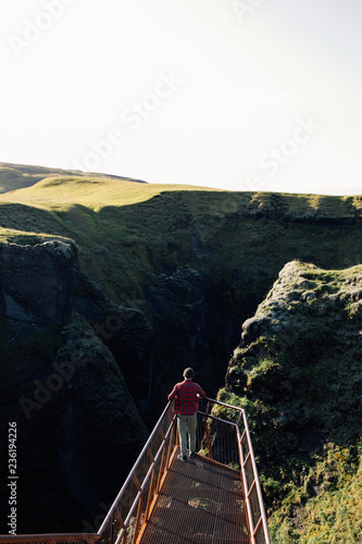 Tourist or traveler man in red checkered shirt and beanie hat stands on viewing point overlooking inspiring epic mountain valley at sunset, new experiences and adrenaline emotions for adventures