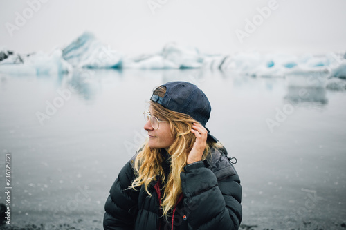 Young pretty blonde woman in puffy down jacket and cap, stand in nordic winter background, tourist looks at icebergs and glaciers floating in ice cold ocean water. Outdoor explore concept