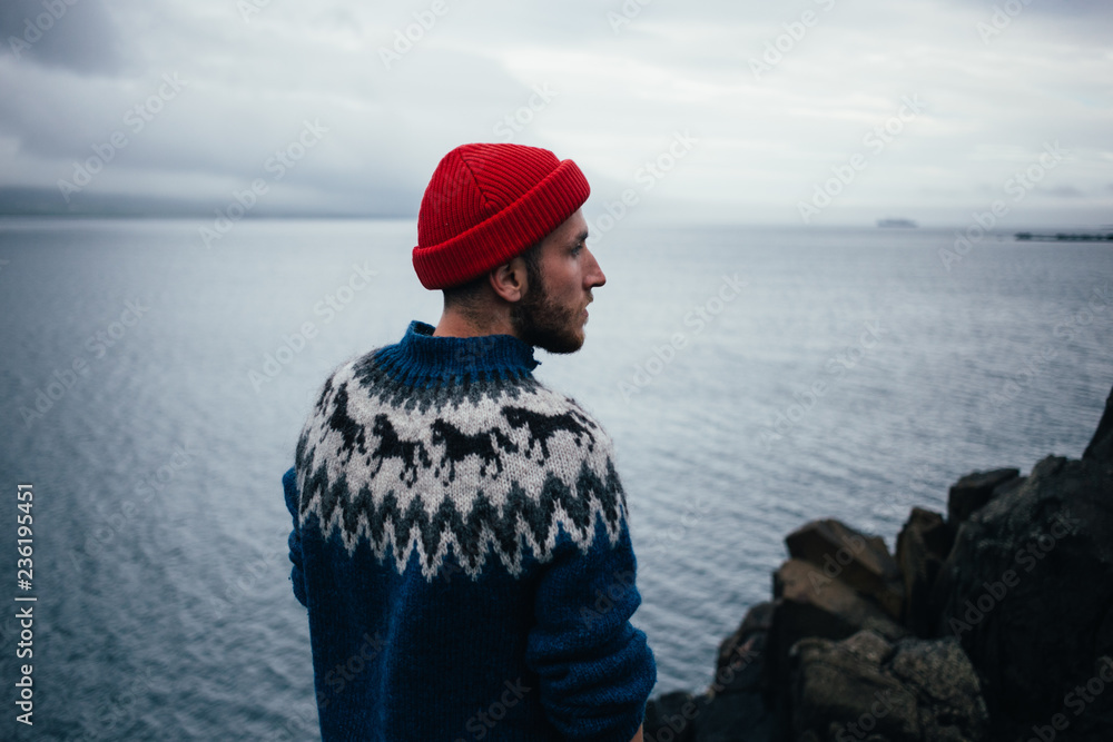 Young handsome man with beard wears traditional authentic blue wool knitted sweater with ornaments and red fisherman or sailor beanie hat, looks to horizon, pensative and thoughtful - obrazy, fototapety, plakaty 
