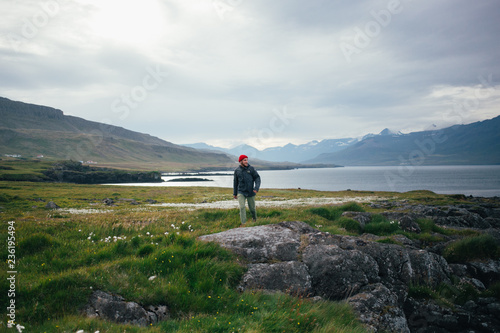 Εκτύπωση καμβά Smiling happy adventurous hiking man, walks in empty spring field or meadow with lush green grass and flowers, epic mountains on background