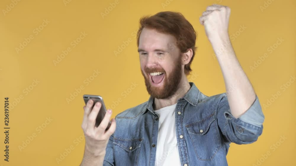Redhead Man Excited for Success while Using Smartphone Isolated on Yellow Background Redhead Man Excited for Success while Using Smartphone Isolated on Yellow Background