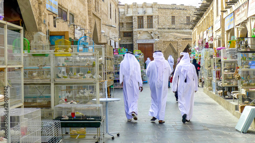 Three men in traditional Qatari dress walk between bird cages and animals for sale in Souq Waqif market in Doha's old town, Qatar