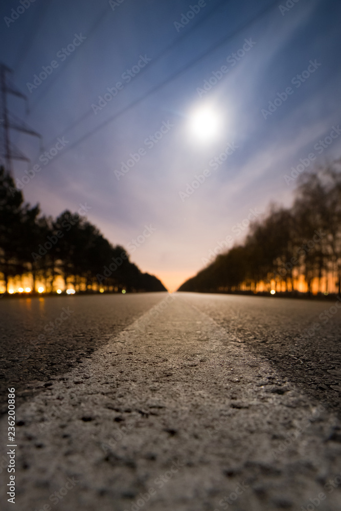 Empty night asphalt road, bright full moon behind clouds and city light behind two rows of trees on both side of road Empty night asphalt road, bright full moon behind clouds and city light behind two rows of trees on both side of road
