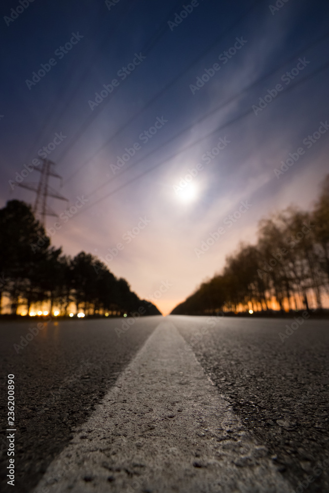 Empty night asphalt road, bright full moon behind clouds and city light behind two rows of trees on both side of road