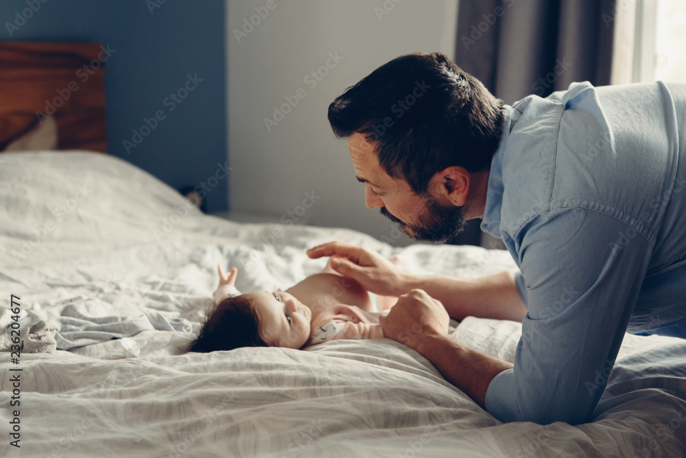 Portrait of middle age Caucasian father talking to newborn baby son daughter. Male man parent smiling to child on bed in bedroom at home. Authentic lifestyle real candid moment. Portrait of middle age Caucasian father talking to newborn baby son daughter. Male man parent smiling to child on bed in bedroom at home. Authentic lifestyle real candid moment.