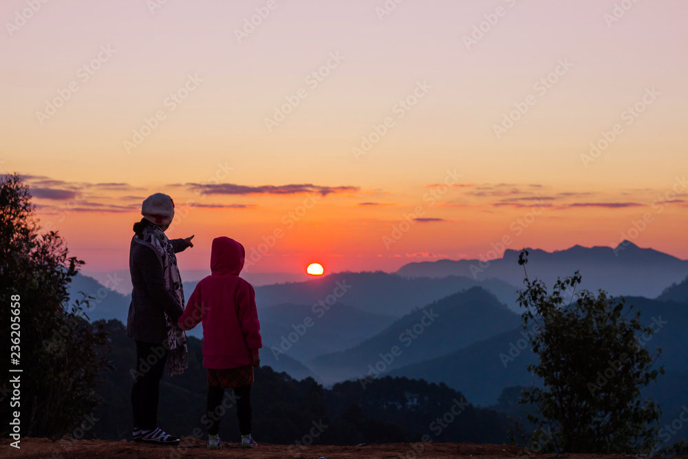 Silhouettes of mother and daughter looking morning sunrise on peak of mountain.