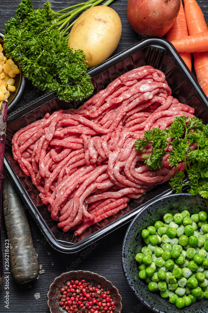 Raw minced meat in container with ingredients for shepherds pie with green peas, yellow corn, carrot, potato, parsley, onion and seasonings, on black background, top view