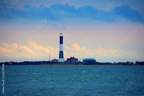Fire Island Lighthouse with beaming light with Long Island Sound, New York, in the foreground with a colorful sky with dark ominous clouds.