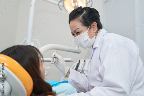 Smiling mature Asian dentist examining teeth of teenage girl
