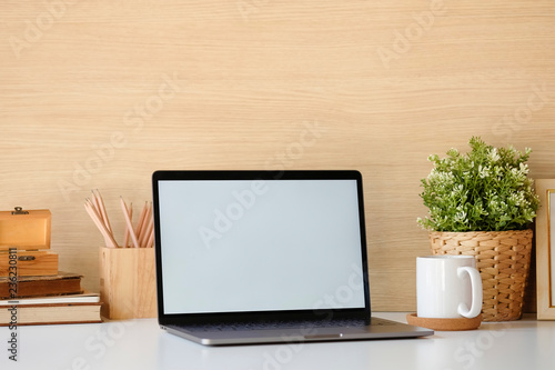 Close-up mockup laptop on white table. books, coffee, pencil. In the background a wood wall and house plants. Empty workplace.