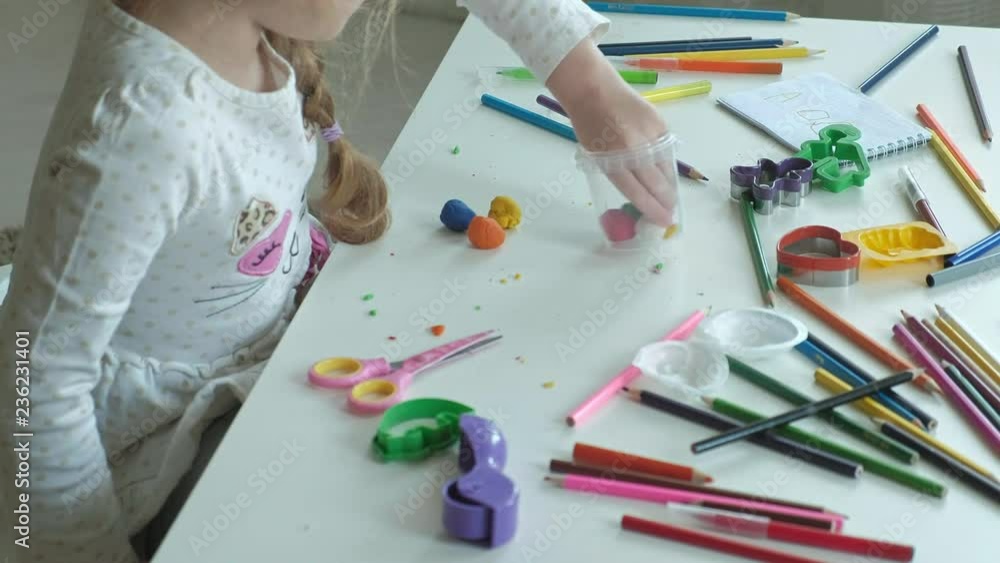 a happy little girl takes out multicolored plasticine from a container, on the desktop are figures and colored pencils, the development of fine motor skills of hands a happy little girl takes out multicolored plasticine from a container, on the desktop are figures and colored pencils, the development of fine motor skills of hands