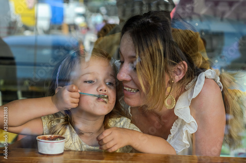 Young mother and daughter eat ice cream in a cafe  happy family  view through the window