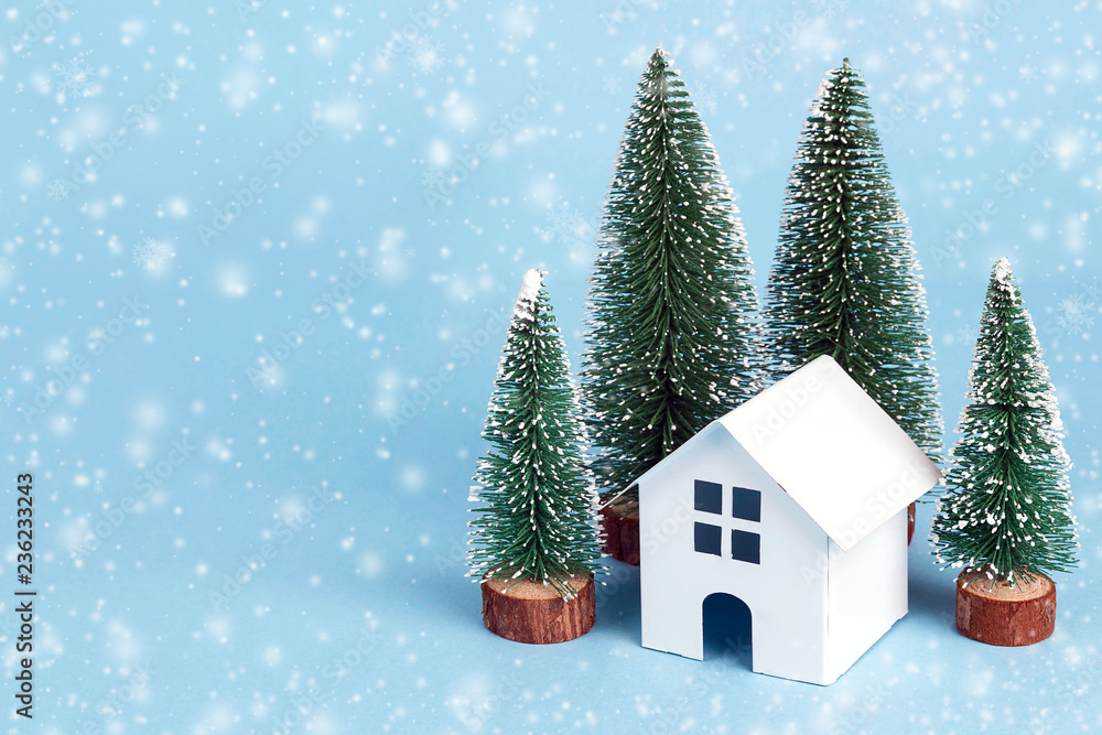 Winter holidays decorative house with fir trees and snowfall on blue background. Copy space for text.