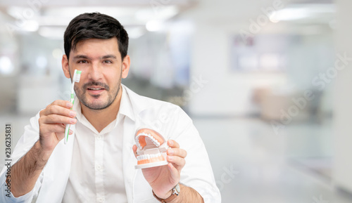 Young male dentist showing toothbrush and denture in dental clinic. Selective focus at the toothbrush. photo