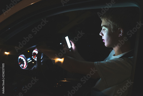 Distracted teenager driving a car with his cell phone in his hand. The light from the screen of the phone is illuminating his face. photo