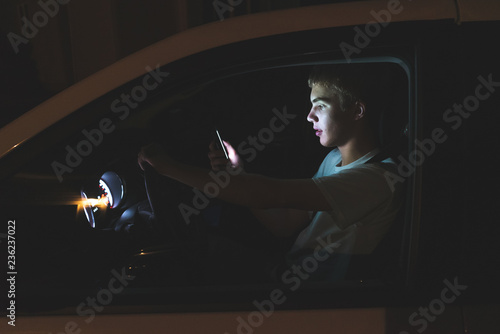 Distracted teenager driving a car with his cell phone in his hand. The light from the screen of the phone is illuminating his face.