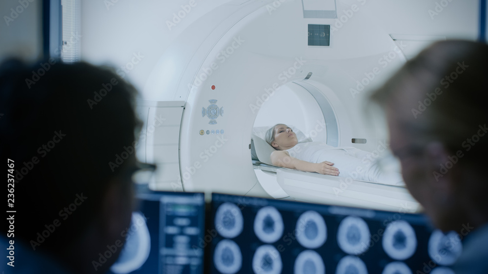 In Control Room Doctor and Radiologist Discuss Diagnosis while Watching Procedure and Monitors Showing Brain Scans Results, In the Background Patient Undergoes MRI or CT Scan Procedure. In Control Room Doctor and Radiologist Discuss Diagnosis while Watching Procedure and Monitors Showing Brain Scans Results, In the Background Patient Undergoes MRI or CT Scan Procedure.