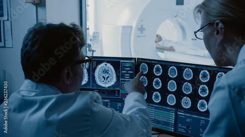In Control Room Doctor and Radiologist Discuss Diagnosis while Watching Procedure and Monitors Showing Brain Scans Results, In the Background Patient Undergoes MRI or CT Scan Procedure.